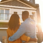 Couple embrace and look up at the outside of their home.